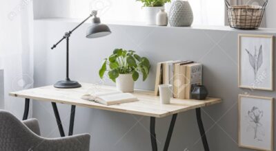 Real photo of wooden desk with fresh plant, black lamp, coffee cup and books standing on half-wall with simple posters
