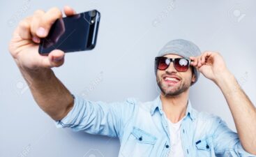 Selfie! Top view of handsome young man in hat and sunglasses making selfie and smiling while standing against grey background